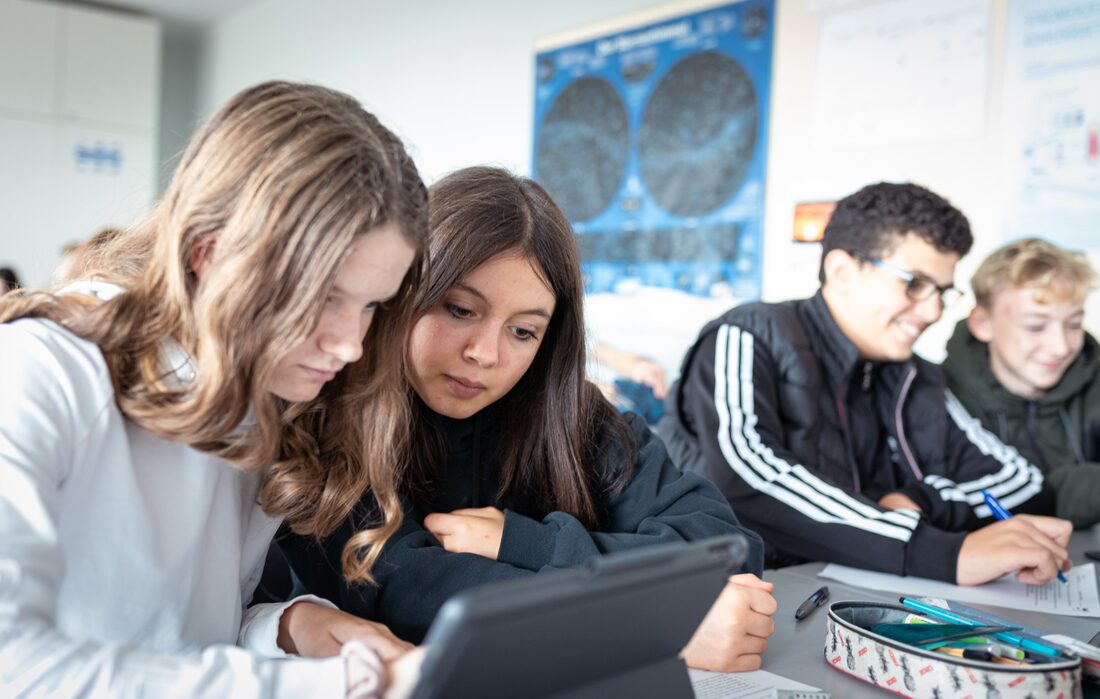 Zwei Schülerinnen arbeiten gemeinsam am iPad, daneben lachen zwei Mitschüler